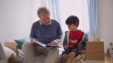 Büyükbaba, sıcak ve eğitici bir aile anını vurgulayarak sıcak, yumuşak ışıklı bir yatak odasında birlikte bir yatakta otururken özenli torununa masal kitabı okuyor.