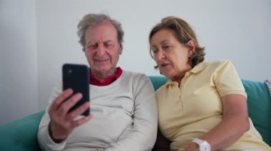 Kanepede birlikte oturan yaşlı bir çift, akıllı telefon ekranına odaklanmış, evlerinin rahatlığında teknolojiyle meşgul oldukları bir an paylaşıyorlar.