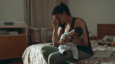 Uyuyan bebeğini kucağında tutan düşünceli bir anne, bir yatakta oturup loş bir yatak odasında anneliğin zorluklarını yansıtıyor.