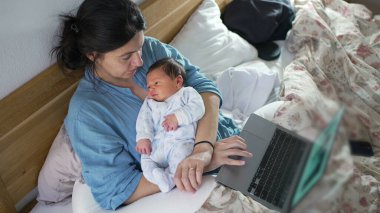 Anne, yeni doğmuş bebeğini yatakta tutarken dizüstü bilgisayarla çalışırken mesleki görevler ve ebeveynlik, modern aile hayatı ve anne bakımı arasındaki dengeyi gösteriyor.
