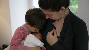 Anne yeni doğan bebeğini emzirirken, büyük kardeş yakından izler, kardeş bağı ve aile birliği anı, yetiştirme ve sevme ortamı