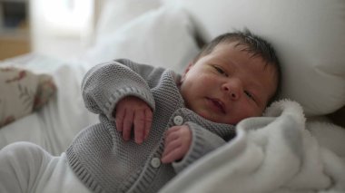 Yeni doğmuş bebek yatağa uzanıyor, gri örgü süveter giyiyor, beyaz pantolon giyiyor, rahat bir battaniyeye sarılmış, küçük eller ve ayaklar görünür, bebek dinleniyor, hayatın ilk anlarını yakalıyor, huzurlu bir sahne.