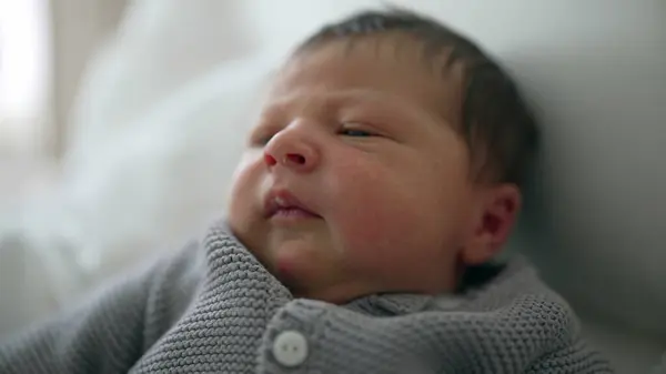 stock image Newborn baby with expressive face, dark hair, and red cheeks, lying on white blanket in gray knit sweater, early days of life, infant facial expression, childhood innocence, cozy environment