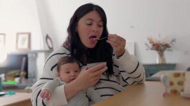 Anne oturma odasında yoğurt yerken bebeğini tutuyor. Samimi ortam ve gündelik an anne ve çocuk arasındaki samimi bağı yansıtıyor.