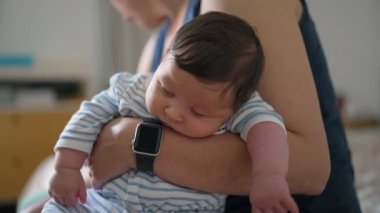 Akıllı saati olan bir annenin kucağında uyuyan bir bebeğin yakın plan fotoğrafı. Kapalı bir ortamda sakin ve huzurlu bir an. Çizgili tulum giymiş bir bebek.