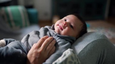 Anne, mutlu bir ifadeyle yukarı bakan bebeğini kucaklıyor. Yumuşak ışık ve rahat ortam oturma odasında sıcak ve besleyici bir atmosfer yaratıyor.