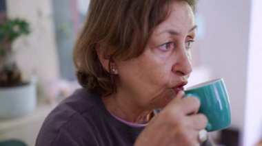 Yeşil fincandan kahve yudumlayan yaşlı bir kadın, barışçıl bir ifadeyle. Basit günlük ritüellerin sıcaklığı ve konforu, sessiz bir tatmin anını yakalıyor.