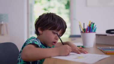 Genç çocuk kalemle çizim yapmaya odaklanmış, renkli kalemler ve sanat malzemeleriyle çevrili bir masada oturuyor, kapalı alanda yaratıcı bir ifade.