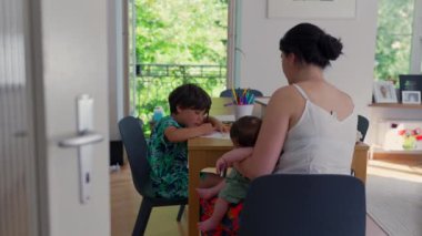 Anne evde eğitim seansı sırasında çocuklarıyla birlikte masada oturur, anne bebeği beşiğinde tutarken gizli eğitim aktiviteleri yapar, huzurlu bir aile anını canlandırır.