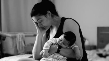 Anne bebeğini yakın tutuyor, görünür bir şekilde yorgun ve derin düşüncelere dalmış bir şekilde, erken anne olmanın duygusal ve fiziksel zorluklarını bir anda yakalayan bir sahne yaratıyor.