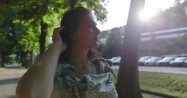 Bir kadın bebekle birlikte ağaçlık bir yolda yürüyor, düşünceli görünüyor, açık şehir manzarası, ağaçların arasından süzülen güneş ışığı, arka planda park edilmiş arabalar.