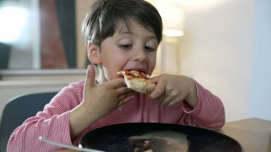 Çocuk yemek masasında bir dilim pizzanın tadını çıkarıyor, yemek sırasında çocukluğun neşesini ve masumiyetini yakalıyor. çocuğun içerik ifadesi ve yeme eylemi