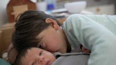 Büyük kardeş ve bebek bir yatakta dinleniyor, rahat bir odada huzurlu ve sevgi dolu bir anı paylaşıyorlar, aralarındaki güçlü bağı ve sevgiyi vurguluyorlar.