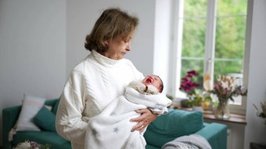 Büyükannesi yeni doğan torununu sıcak bir battaniyeye sardı, parlak bir ev ortamında, aile sevgisinin, sıcaklığın ve nesiller arası bağın hassas bir anını yakalıyor.