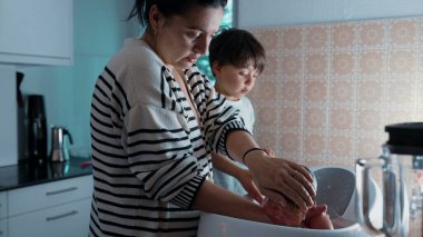 Anne, küçük bir küvette yeni doğmuş bir bebeği dikkatlice yıkıyor, büyük kardeş izliyor, aile bakımını ve bağ kurmayı vurguluyor. Bir bebeği besleyen ve koruyan anlar.