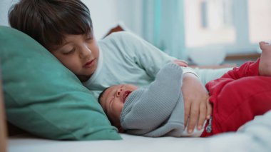 Genç bir çocuk yeni doğmuş kardeşini kucaklarken yatakta uzanmış, güzel bir kardeşlik bağı kuruyor.