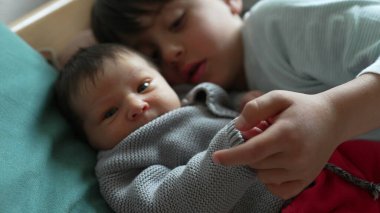 Büyük kardeş bebeğin elini nazikçe tutuyor, rahat bir evde bir yatakta uzanıyor, iki çocuk arasında yakın ve sevgi dolu bir bağ kuruyor.