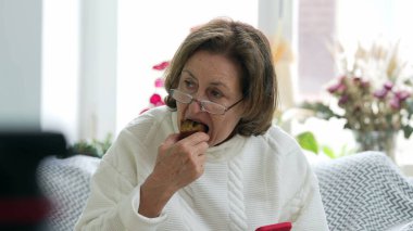 Kanepede abur cubur yiyen, gözlük takan ve beyaz kazak giyen yaşlı bir kadın, huzur, rahatlık ve günlük aktiviteleri bir ev ortamında ön plana çıkarıyor.