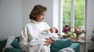 Büyükannesi yeni doğan torununu sıcak bir battaniyeye sardı, parlak bir ev ortamında, aile sevgisinin, sıcaklığın ve nesiller arası bağın hassas bir anını yakalıyor.