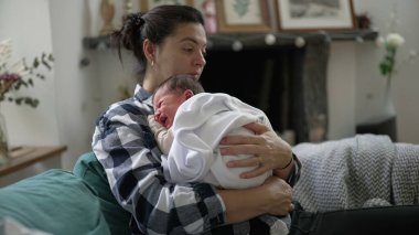 Anne, yeni doğmuş bebeğini bir battaniyeye sarılmış bir şekilde teselli ederken, bir ev ortamında kanepede oturup, aralarındaki hassas ve besleyici bağı vurguluyor.