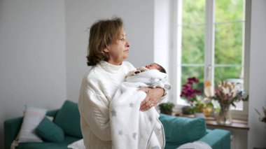 Büyükannesi yeni doğan torununu sıcak bir battaniyeye sardı, parlak bir ev ortamında, aile sevgisinin, sıcaklığın ve nesiller arası bağın hassas bir anını yakalıyor.