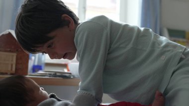 Büyük kardeş ve bebek bir yatakta sevgi dolu, güneşli, oyuncaklarla dolu sıcak bir odada hassas ve neşeli anlar paylaşıyorlar, güçlü bir kardeş bağı ve neşeli bir aile ortamı sergiliyorlar.