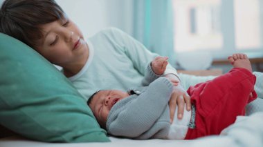 Genç bir çocuk yeni doğmuş kardeşini kucaklarken yatakta uzanmış, güzel bir kardeşlik bağı kuruyor.