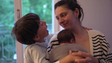 Anne, yeni doğan ve daha büyük olan çocuğunu kucaklıyor, o da onu yanağından öpüyor. derin aile sevgisi ve sevgi, anne ve çocuklar arasındaki bağı vurguluyor.
