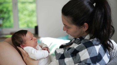 Anne onu kollarında tutarken sevgi dolu bir şekilde esneyen bebeğine bakıyor. Sıcak bir evde gün ışığı, güçlü bir duygusal bağ ve aile bağıyla geçen samimi bir an.
