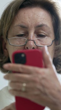 Gözlük takan yaşlı bir kadın, akıllı telefonuna dikkatle bakıyor, modern teknoloji kullanımının yakın çekim anı ve ev ortamında konsantrasyon