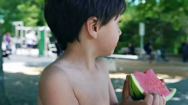 stock image Young boy focused on eating a juicy watermelon slice, enjoying a sunny day in the park, shirtless with a relaxed expression, healthy summer snack, refreshing and hydrating, outdoor fun