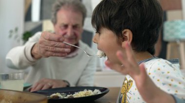 Yaşlı bir adam, bir çocuğun kaşıkla yemesine yardım ediyor, bir ev ortamındaki yemek sırasında etkileşime yakın çekim yapıyor, nesiller boyunca sevgi dolu bir aile ortamında bakıma ve bakıma odaklanıyor.