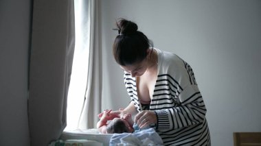 Anne, yeni doğmuş bebeğini pencerenin kenarındaki değişen masada nazikçe bekliyor. Huzurlu bir evde sevgi ve ilgi dolu bir an sergiliyor.