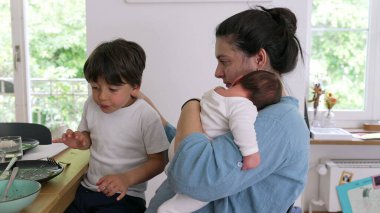 Anne yemek masasında büyük bir çocuğun yanında otururken bebeği tutuyor. Sahne, sıcak bir ev ortamında ebeveynliğin çoklu görev ve besleyici yönlerini yansıtıyor.