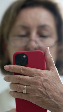 Gözlük takan yaşlı bir kadın, akıllı telefonuna dikkatle bakıyor, modern teknoloji kullanımının yakın çekim anı ve ev ortamında konsantrasyon