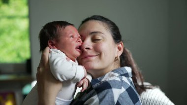 Gülümseyen anne, esneyen bebeğini yüzüne yakın tutuyor. Sevecen bir sevinç ve bağlantı anını paylaşıyor.