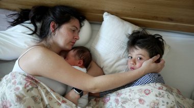 Anne ve iki çocuğu yatakta, yeni doğmuş bebek annesinin kollarında, küçük çocuk uyanık ve neşeli, sabah aile zamanı, sıcak ve sevgi dolu, samimi anlar, sıcak ev ortamı