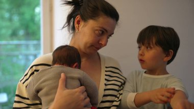 Anne, büyük çocuğunu azarlarken yeni doğmuş bebeğini bir inançsızlık ifadesiyle kucaklıyor. Aile ilişkilerinin dinamiği ve annenin özenli doğası.