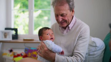 Yeni doğmuş bir bebeği kucaklayan yaşlı bir adam, her ikisi de pencerenin yumuşak doğal ışığıyla sıcak bir ev ortamında şefkatli bakışlarını paylaşıyor, yürek ısıtan bir bağ ve sevgi anı sergiliyor.