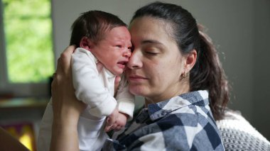 Gülümseyen anne, esneyen bebeğini yüzüne yakın tutuyor. Sevecen bir sevinç ve bağlantı anını paylaşıyor.