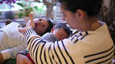 Anne yeni doğan bebeğini nazikçe tutarken, büyük kardeş eğilip, bir aile birlikteliği ve sevgi anı sergiliyor. Sahne, aralarındaki bağları yakalar.