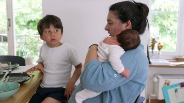 Anne yemek masasında büyük bir çocuğun yanında otururken bebeği tutuyor. Sahne, sıcak bir ev ortamında ebeveynliğin çoklu görev ve besleyici yönlerini yansıtıyor.