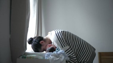 Anne, yeni doğmuş bebeğini pencerenin kenarındaki değişen masada nazikçe bekliyor. Huzurlu bir evde sevgi ve ilgi dolu bir an sergiliyor.