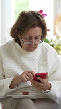 Gözlüklü yaşlı bir kadın, bir parça kek yerken akıllı telefonuna odaklanmış, modern bir teknolojik kullanım anı ve parlak bir ev ortamında sıradan bir akşam yemeği.
