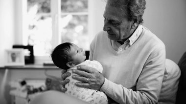 Büyükbaba, siyah-beyaz fotoğrafta yeni doğan bebeği kucaklıyor, hassas ve samimi bir an, aile sevgisi, nesiller arası bağ, rahat yuva kurma, yaşlılar ve bebekler arası bağ, sonsuz sevgi.