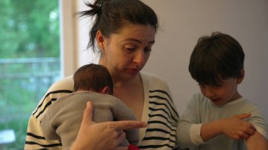 Anne, büyük çocuğunu azarlarken yeni doğmuş bebeğini bir inançsızlık ifadesiyle kucaklıyor. Aile ilişkilerinin dinamiği ve annenin özenli doğası.