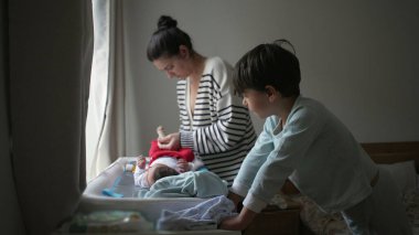 Anne ve küçük kardeş yeni doğmuş bir bebeğe bakmakla ilgilenirler. Değişen bir masada sıcak bir aile etkileşimi ve yuva ortamında bağ kurma anını vurgularlar.