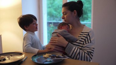 Anne yeni bebeğini kucağında tutarken, büyük kardeş sevgiyle bebekle etkileşime giriyor. Yemek masasındaki iç açıcı sahne aile bağlarının özünü yakalar.