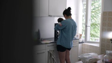 Anne 'nin bebeğini kucağında tutarken, parlak bir mutfakta yemek hazırlarken, birden fazla işi olan ve ebeveynliği günlük ev işleri, sevgi ve bakım ile dengeleyen,