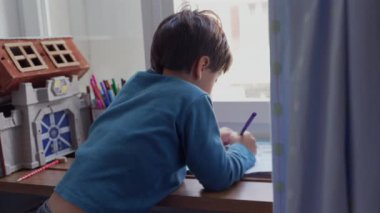 Pencere kenarına yaslanmış, resim yapmaya odaklanmış, etrafı renkli kalemler ve oyuncaklarla çevrili genç bir çocuk. Bir çocuğun konsantrasyonu ve yaratıcılığı bir sanat aktivitesinde özümsenmiştir.